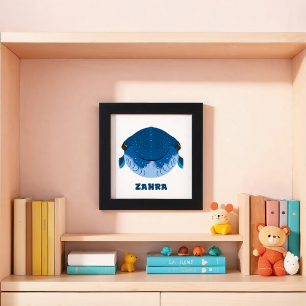 Illustration d’une baleine souriante et le prénom Zahra en dessous dans un cadre noir, posé dans une chambre d’enfant.